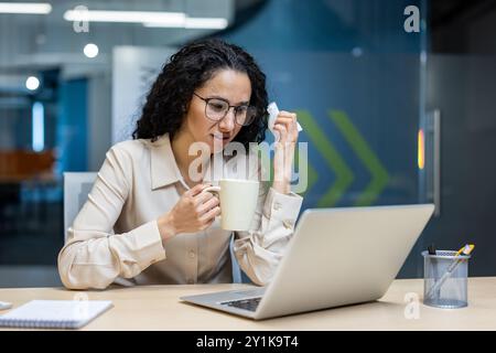 Femme d'affaires se sentant mal au lieu de travail boit des médicaments chauds et tient une serviette. Assise à son bureau avec son ordinateur portable, elle se concentre sur son travail malgré sa maladie. Engagement de l'environnement de bureau en cas de maladie. Banque D'Images