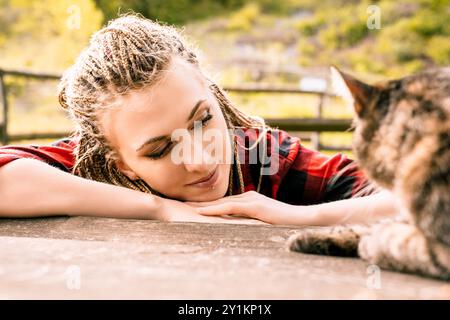 Jeune femme avec des tresses repose sa tête sur ses bras sur une surface en bois, profitant d'un moment de paix avec son chat dans la nature Banque D'Images