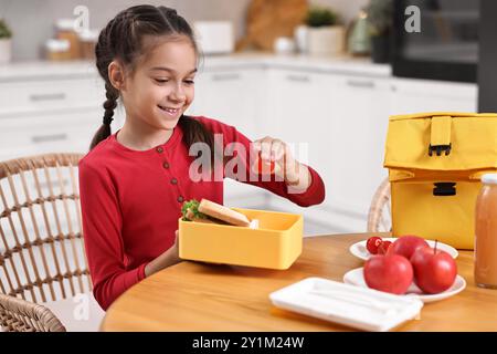Fille mignonne mettant la tomate dans la boîte à lunch à la table en bois dans la cuisine. Préparation à l'école Banque D'Images
