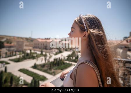 Une jeune femme aux longs cheveux bruns se dresse sur un balcon donnant sur un paysage urbain tentaculaire avec une végétation luxuriante. Elle regarde la vue lointaine, ses yeux c Banque D'Images