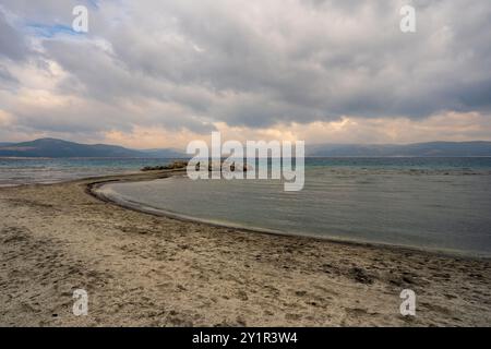 Une scène tranquille au lac Salda, en Turquie, avec une plage de sable qui se courbe au loin, des eaux claires et des montagnes sous un ciel nuageux. Banque D'Images