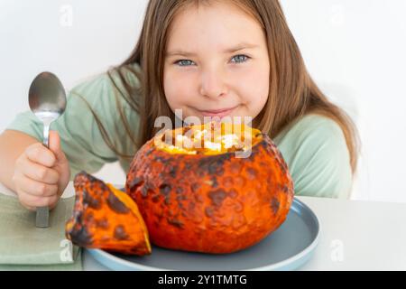 Petite adolescente mangeant de la soupe de citrouille servie dans de la citrouille cuite au four. Fille en t-shirt vert et plat d'halloween d'automne sur fond blanc. Pho de haute qualité Banque D'Images