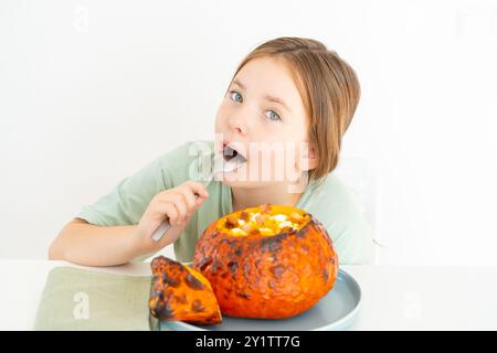 Petite adolescente mangeant de la soupe de citrouille servie dans de la citrouille cuite au four. Fille en t-shirt vert et plat d'halloween d'automne sur fond blanc. Pho de haute qualité Banque D'Images