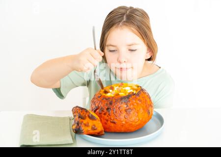 Petite adolescente mangeant de la soupe de citrouille servie dans de la citrouille cuite au four. Fille en t-shirt vert et plat d'halloween d'automne sur fond blanc. Pho de haute qualité Banque D'Images