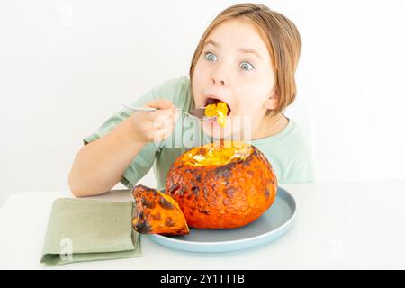 Petite adolescente mangeant de la soupe de citrouille servie dans de la citrouille cuite au four. Fille en t-shirt vert et plat d'halloween d'automne sur fond blanc. Pho de haute qualité Banque D'Images