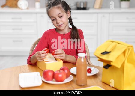 Fille mignonne mettant des tomates dans la boîte à lunch à la table en bois dans la cuisine. Préparation à l'école Banque D'Images