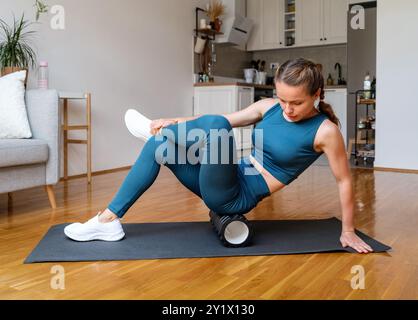 Femme athlétique fait une séance d'entraînement à la maison, en utilisant un rouleau en mousse pour libérer les muscles des jambes Banque D'Images