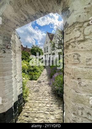 Bruges, Flandres, Belgique - 22 juin 2024 : Ten Wijngaerde Béguinage jardin privé avec des fleurs sur le côté NW sous nuages bleus, regardant à travers l'arche Banque D'Images