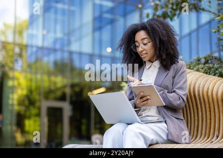 Femme d'affaires afro-américaine participe à un cours vidéo en ligne à l'extérieur. Elle communique avec l'instructeur via un ordinateur portable, prend des notes dans les utilisations de l'ordinateur portable pour les appels vidéo, améliorant ainsi ses compétences professionnelles Banque D'Images