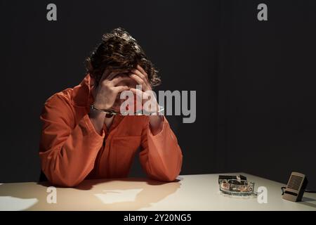 Individu en combinaison orange assis à table avec les mains retenues, la tête reposant sur les mains comme si dans la pensée profonde ou l'inquiétude. Les menottes visibles soulignent la gravité du scénario Banque D'Images