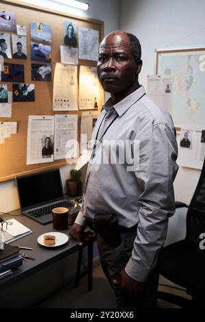 Homme afro-américain âgé debout au bureau, examinant des photos de scène de crime et des documents à bord, avec une expression sérieuse, regardant la caméra Banque D'Images
