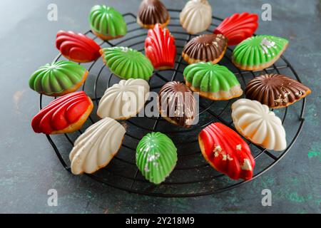 Biscuits madeleine glacés assortis sur grille de pâtisserie ronde sur fond sombre. Biscuits madeleine aux baies, chocolat, menthe et crème au beurre sur un noir Banque D'Images