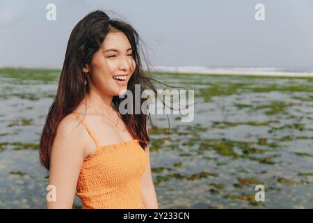 Heureuse jeune femme asiatique portant une robe orange se moque de la caméra avec l'océan en arrière-plan. Banque D'Images