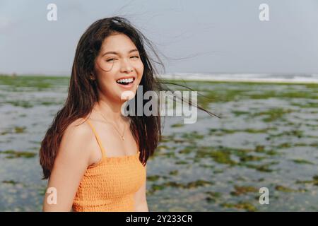 Heureuse jeune femme asiatique portant une robe orange se moque de la caméra avec l'océan en arrière-plan. Banque D'Images