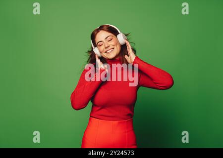 Jeune femme dans des vêtements décontractés se tient en toute confiance sur un fond vert vif, portant un casque antibruit et écoutant de la musique. Femme enjo Banque D'Images
