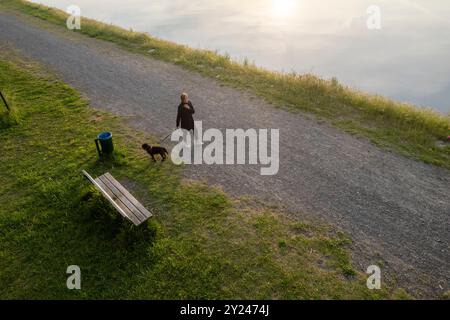 Femme prend une promenade paisible le soir avec son chien le long d'un chemin au bord d'un lac serein, le soleil couchant jetant une lueur chaude sur l'eau Banque D'Images