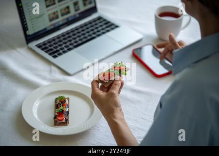 Femme a sandwich de pain croustillant de seigle avec du fromage à la crème et du thé à table. Bouchée rapide, addiction aux gadgets Banque D'Images