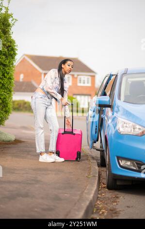 Femme indienne dans une tenue en denim élégante avec une valise rose, voyageant à l'aéroport en voiture et en bus, vérifiant son téléphone en attendant. Banque D'Images