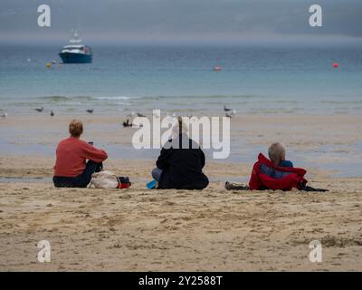 Trois personnes assises sur la plage, jour de pluie, port de St Ives, St Ives, Cornwall, Angleterre, Royaume-Uni, GB. Banque D'Images
