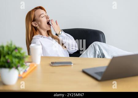 Femme endormie vêtue d'une chemise assise à son lieu de travail au bureau, bâillant Banque D'Images