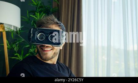 Homme drôle sourit tout en utilisant un casque VR dans un salon moderne Banque D'Images