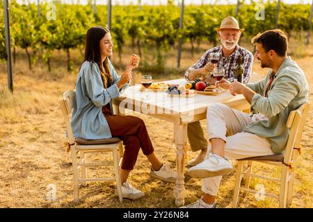 Trois amis se réunissent autour d'une table rustique dans un vignoble, dégustant du vin, des fruits frais, et la compagnie de l'autre sur une journée ensoleillée remplie de joie et de lau Banque D'Images