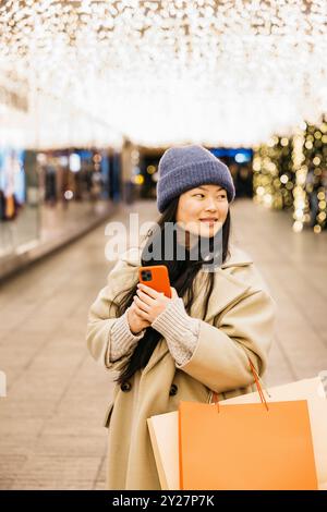 Portrait d'une jeune femme asiatique tenant des sacs à provisions, et un téléphone, pendant une journée de shopping de noël. Banque D'Images