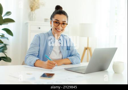 Photo d'une jolie femme confiante. Femme arabe ou indienne réussie dans une chemise bleue, travaillant sur l'ordinateur portable, développant un nouveau projet numérique, planifiant une stratégie marketing, regardant et souriant à la caméra Banque D'Images
