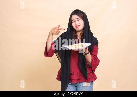 Belle femme asiatique mature portant une chemise rouge portant un hijab avec une expression sullen pleurant pointant vers le bas et portant une assiette contenant du chopsti Banque D'Images