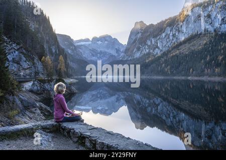 Une femme faisant du yoga au bord du lac. Position Lotus (Padmasana) . Vorderer Gosausee en automne avec vue sur les montagnes de Dachstein. Le Gosaukamm sur le Banque D'Images