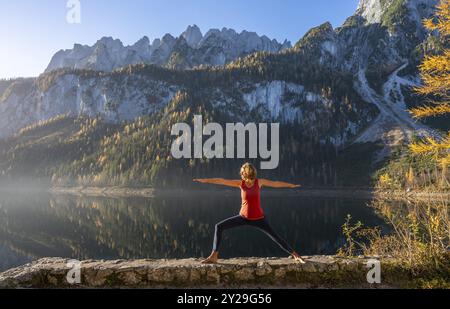 Une femme faisant du yoga au bord du lac. Guerrier 2 (Virabhadrasana) . Vorderer Gosausee en automne avec vue sur la chaîne de montagnes du Dachstein. Le Gosaukamm sur Banque D'Images