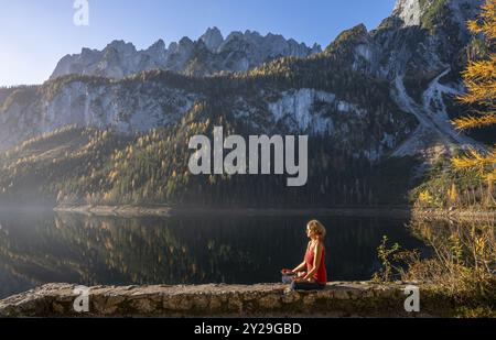 Une femme faisant du yoga au bord du lac. Position Lotus (Padmasana) . Vorderer Gosausee en automne avec vue sur les montagnes de Dachstein. Le Gosaukamm sur le Banque D'Images