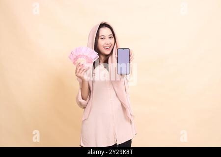 Fille asiatique souriant heureux à la caméra transportant de l'argent rupiah et tenant l'écran de gadget de téléphone portable devant elle. beau jeune pour la finance, t Banque D'Images