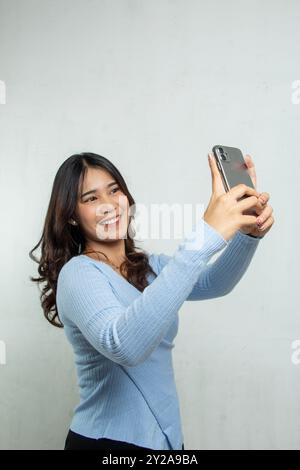 Belle fille asiatique dans le costume haut bleu clair décontracté prenant selfie avec le téléphone portable et souriant joyeusement, femme adulte debout utilisation dans la publicité Banque D'Images