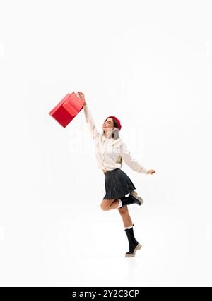 Jeune femme saute joyeusement dans l'air, tenant des sacs à provisions rouges vibrants sur fond blanc de studio. Une virée shopping réussie Banque D'Images