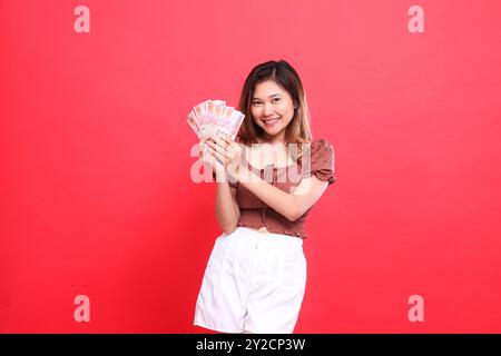 Joyeuse expression de femme indonésienne tenant de la monnaie rupiah dans les deux mains, portant une blouse brune sur un fond rouge. pour la transaction, technologie a. Banque D'Images