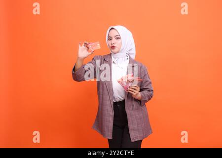 Jeune femme indonésienne de 20 ans portant des vêtements de bureau hijab tenant une carte de crédit de débit finissent par embrasser la pose et l'argent à côté isolé sur le pastel Uni Banque D'Images