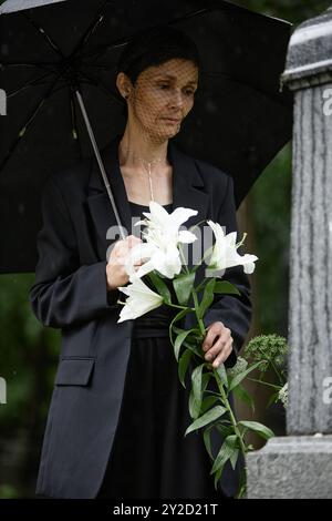 Plan vertical d'une femme âgée en costume funéraire tenant des lis blancs visitant la tombe d'un être cher disparu tout en se cachant de la pluie sous parapluie au cimetière Banque D'Images