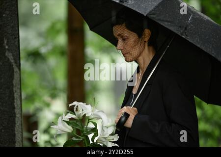 Vue de côté d'une femme âgée en deuil voile pleurant sur la perte d'un être cher au cimetière tenant des lis blancs avant de placer des fleurs à la tombe tout en se cachant de la pluie sous parapluie noir, espace de copie Banque D'Images