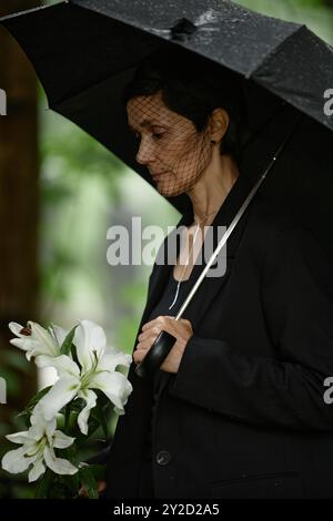 Plan vertical d'une femme âgée en costume noir et voile de deuil sous parapluie pleurant de la mort d'un être cher au cimetière, tenant des lis blancs mouillés par la pluie avant de placer des fleurs à la tombe Banque D'Images