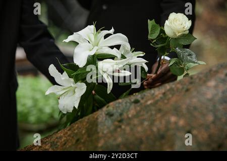 Gros plan sur la pluie lys blancs humides et rose dans les mains de deux personnes méconnaissables plaçant des fleurs sur le monument commémoratif en pierre pour commémorer un être cher décédé au cimetière, espace copie Banque D'Images