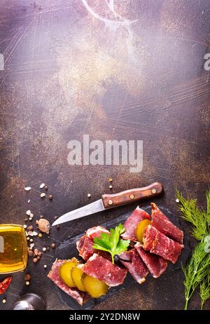 Tranches de pastrami séchées, viande de bœuf aux herbes et concombre mariné sur tableau noir. Banque D'Images