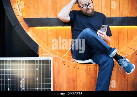 Homme barbu utilisant un smartphone connecté à un panneau solaire photovoltaïque. Intégration de l'énergie renouvelable durable dans la vie quotidienne, montrant l'utilisation pratique de l'énergie solaire. Vue rognée. Concentrez-vous sur le téléphone. Banque D'Images