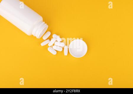 Un flacon de médicament blanc versant des pilules blanches sur un fond jaune. L'image est prise d'en haut, adapté aux visuels de santé et pharmaceutiques Banque D'Images