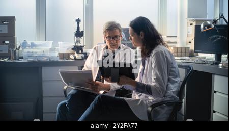 Deux scientifiques divers en blouse de laboratoire discutent de la recherche scientifique, regardent des articles et utilisent une tablette. Les paléontologues ou archéologues travaillent dans un laboratoire archéologique avec des équipements modernes. Banque D'Images