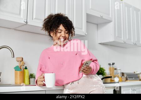 Femme souriante se détend avec un café tout en utilisant son téléphone dans une cuisine lumineuse. Banque D'Images