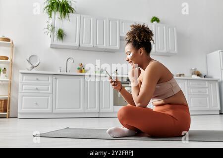 La femme est assise confortablement à la maison, souriante tout en utilisant son smartphone sur un tapis. Banque D'Images