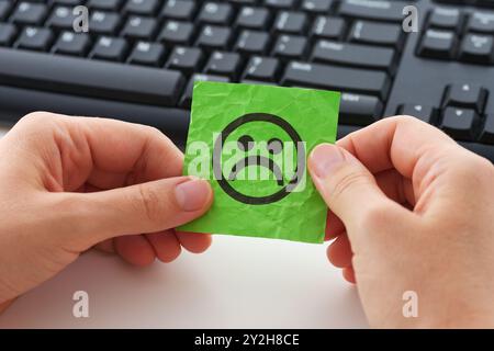 Une femme d'affaires dans un bureau assise derrière un ordinateur et tenant un morceau de papier vert avec un visage triste dessus dans ses mains. Stress sur le lieu de travail. Banque D'Images