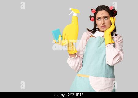 Portrait de jeune femme au foyer fatiguée dans un tablier avec éponge et détergent sur fond gris Banque D'Images