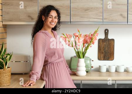 Belle jeune femme afro-américaine heureuse avec vase de bouquet de fleurs de gladiolus rose dans la cuisine à la maison Banque D'Images